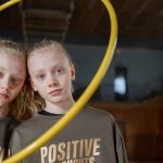 Twin girls in sportswear indoors with a hula hoop expressing positivity. Perfect for family and sports themes.